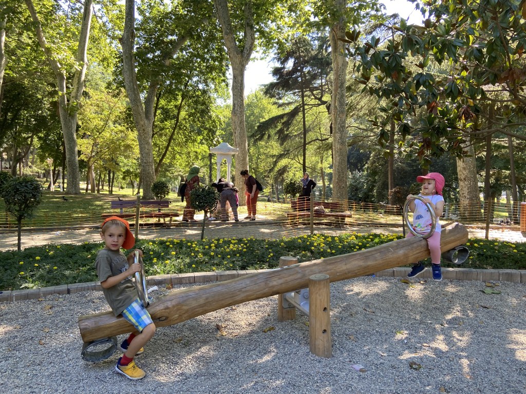 Two children playing on a wooden seesaw in a park, surrounded by greenery and other people enjoying the area.