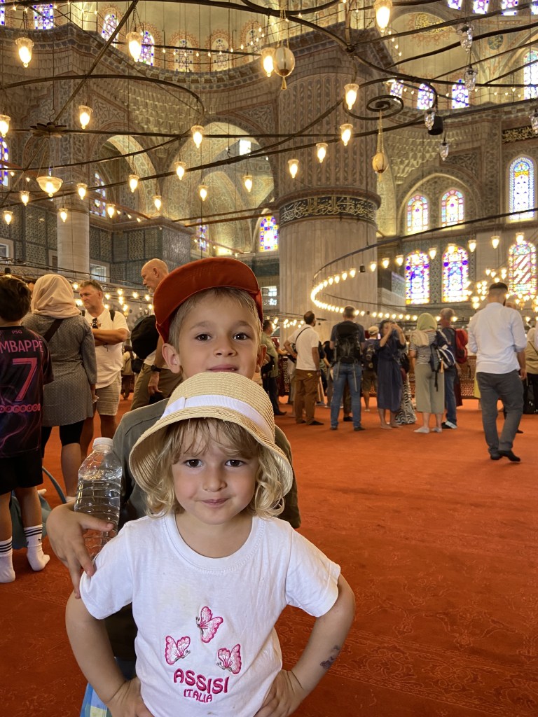 Two children posing together inside a mosque with decorative lights and stained glass windows in the background.