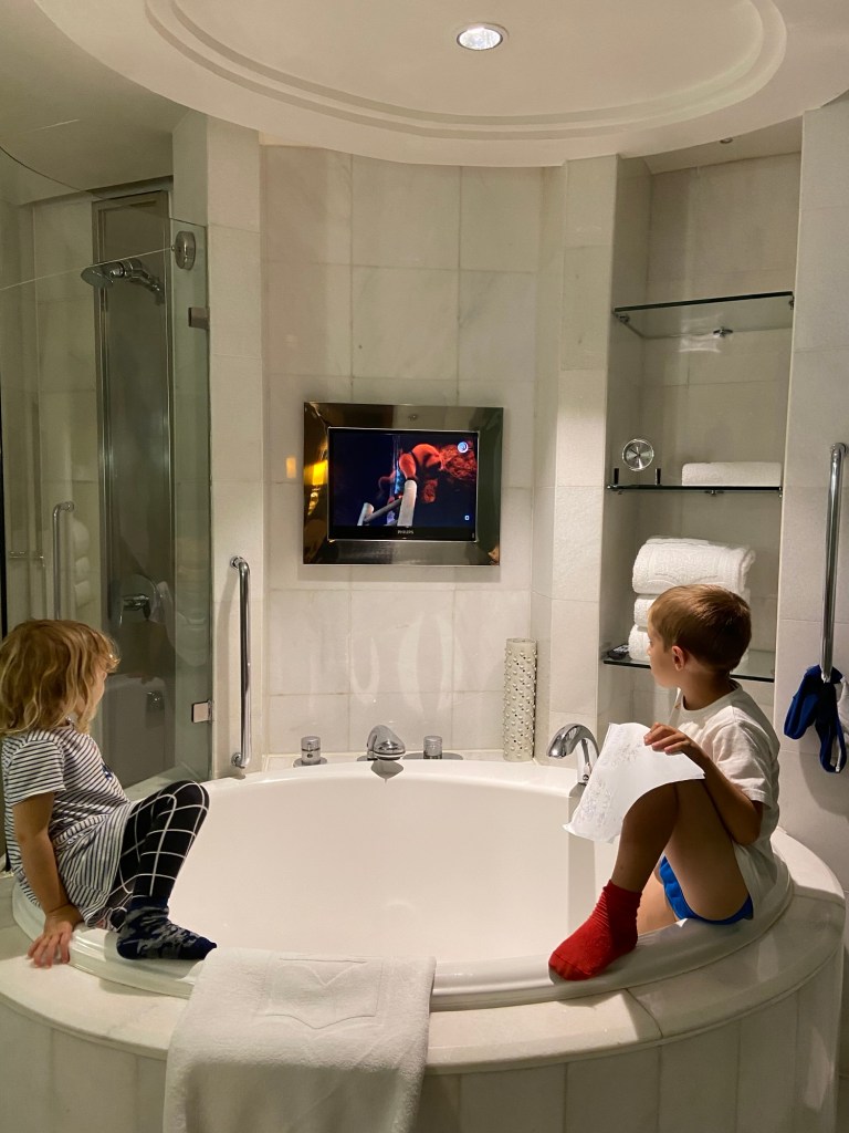 Two children sitting on the edge of a round bathtub, watching a television mounted on the wall in a modern bathroom.