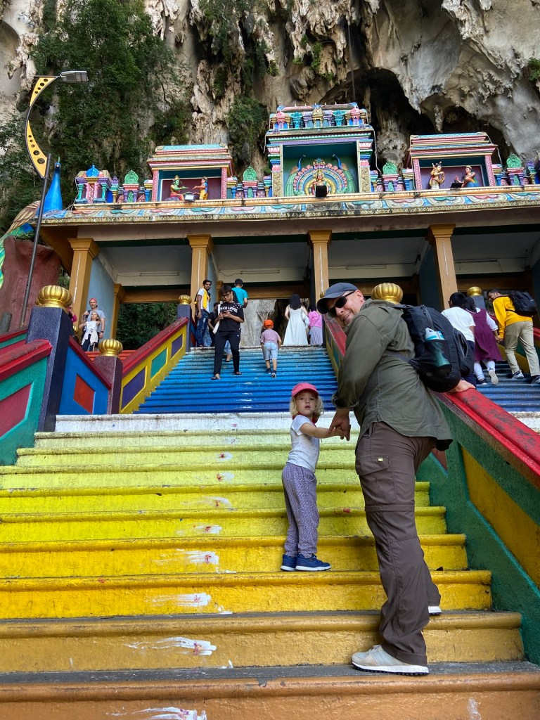 A parent and child standing on colorful steps at a temple, with people climbing the stairs in the background and a lush cliff above.
