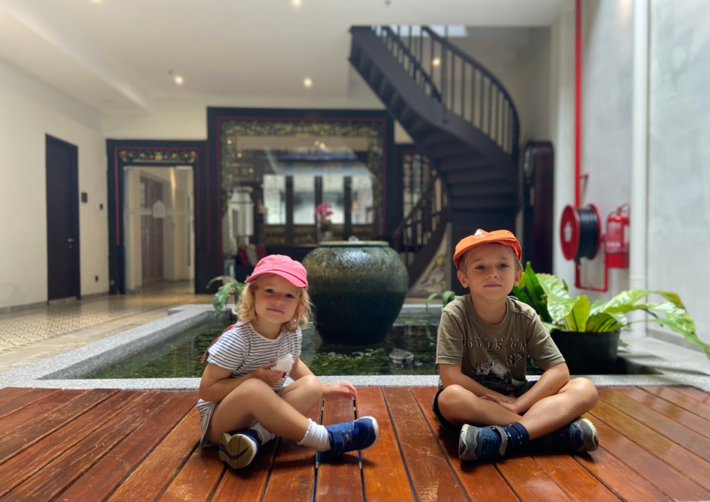 Two children sitting on a wooden floor in a modern indoor space with a water feature and plants in the background.