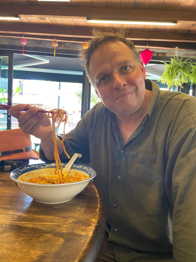 A man sitting at a wooden table in a restaurant, holding chopsticks with noodles, looking at the camera with a slight smile. A bowl of noodles with vegetables is placed in front of him.