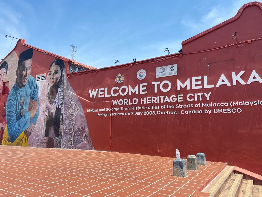 Murals on a red wall welcoming visitors to Melaka, Malaysia, with text stating 'WELCOME TO MELAKA WORLD HERITAGE CITY'.