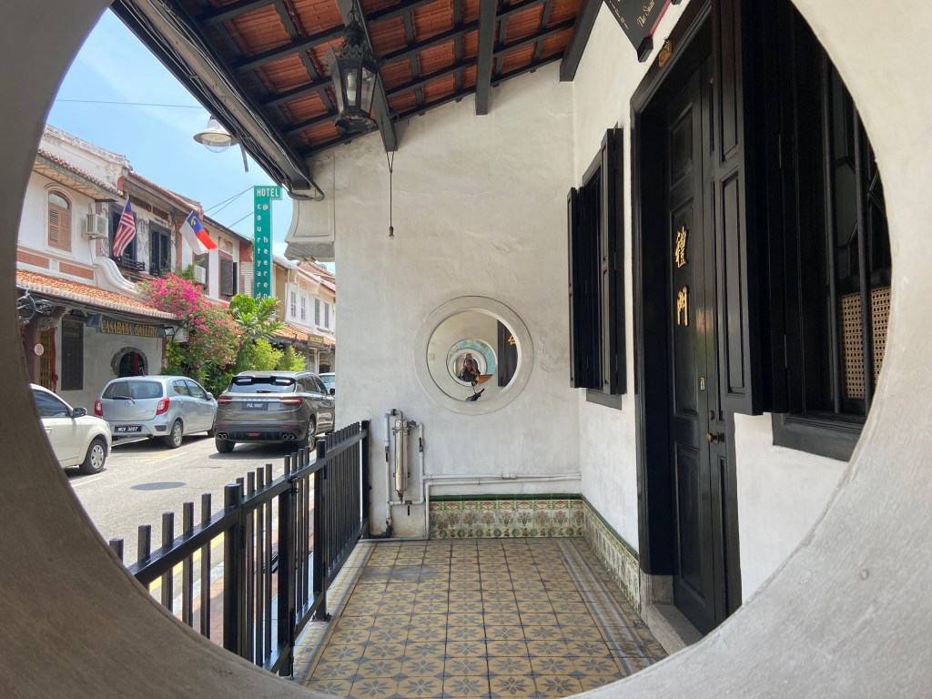 A view through a circular archway leading to a traditional building with black doors and large windows, showcasing a tiled floor and decorative wall patterns. In the background, a street scene features colorful buildings, parked cars, and plants.